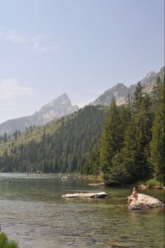 a person sitting on a rock in the middle of a body of water with mountains in the background