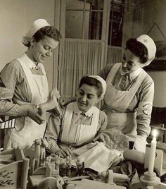 three women in aprons standing around a table with bottles and candles on it, all smiling