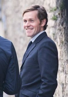 a man in a suit and tie standing next to a stone wall smiling at the camera