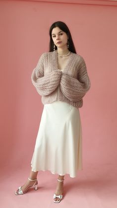 a woman standing in front of a pink wall wearing a white skirt and grey sweater