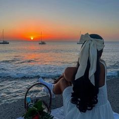 a woman sitting on top of a beach next to the ocean at sunset with boats in the background