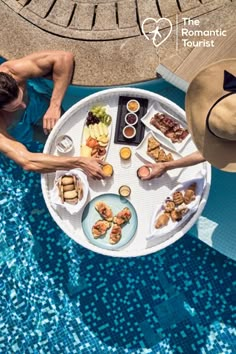 two people sitting at a table with food on it near a swimming pool in the sun