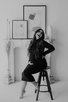 black and white photo of woman sitting on stool in front of fireplace with framed artwork