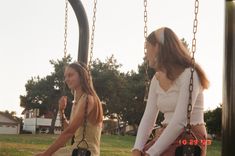 two young women sitting on swings in a park, one holding onto the other's hand