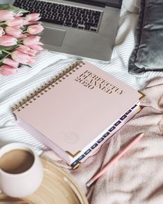 a pink notebook sitting on top of a bed next to a coffee cup and laptop