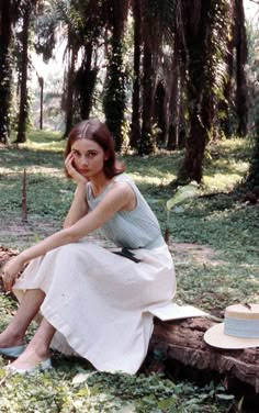 a woman is sitting on a log in the woods wearing a dress and hat with her hand under her chin