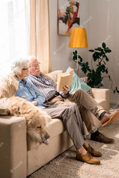 an elderly man and woman sitting on a couch reading a book with their dog in front of them