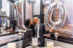 a man and woman kissing in front of a cake on a table with giant balloons