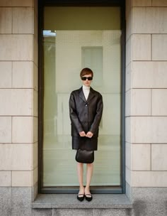 a woman standing in front of a window wearing a black jacket and skirt with sunglasses on her head