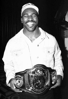 a black and white photo of a smiling man holding a wrestling belt in his hands