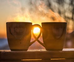 two coffee mugs sitting on top of a wooden table with the sun setting behind them