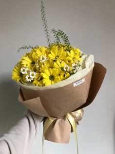 a person holding a bouquet of yellow and white flowers in brown paper wrapped around it