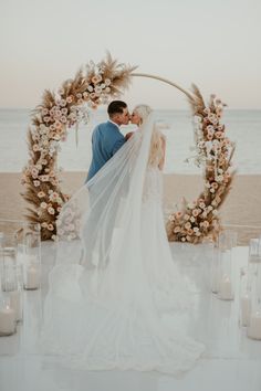 a bride and groom kissing in front of an arch with candles on it at the beach