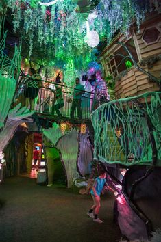 people are walking around in an indoor area with plants and trees on the walls, lights hanging from the ceiling