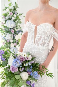 a woman in a wedding dress holding a bouquet of purple and white flowers with greenery behind her