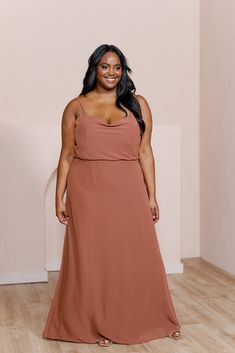 a woman in a long brown dress standing on a wooden floor with her hands behind her back