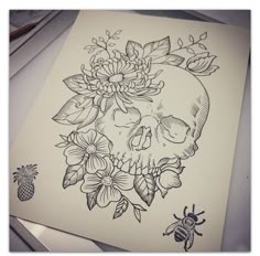 a drawing of a skull with flowers and a bee on it's side, sitting next to a piece of paper