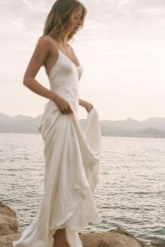 a woman in a white dress standing on rocks by the water with her hands behind her back