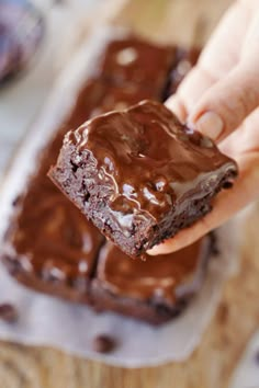 a person holding a piece of chocolate brownie on top of a wooden cutting board