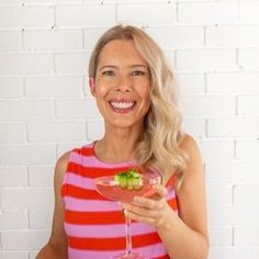 a woman holding a wine glass filled with fruit and veggies in front of a brick wall