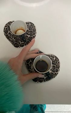 two people holding coffee cups and saucers on top of a white table next to each other