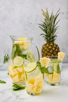 two glasses filled with lemonade and lime slices next to a pitcher of pineapple