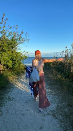 a woman walking down a dirt road towards the ocean with her arms out to the side