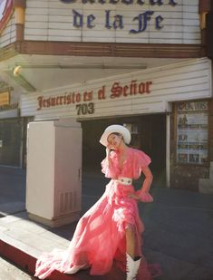 a woman in a pink dress and cowboy hat standing on the side of a street