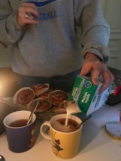 a person pouring milk into some food on a table next to cups and spoons