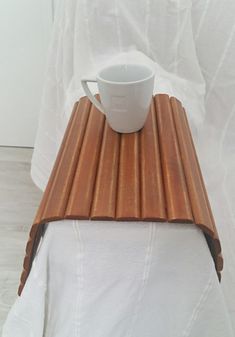 a white cup sitting on top of a wooden table next to a white cloth covered bed