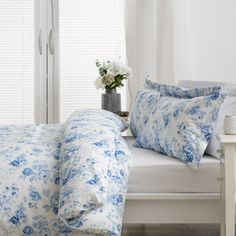 a bed with blue and white comforters in front of a window next to a vase filled with flowers