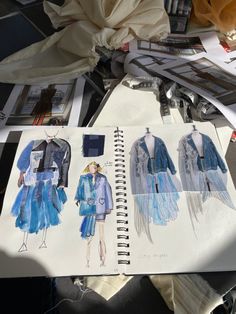 an open notebook with drawings of dresses and jackets on top of it next to papers