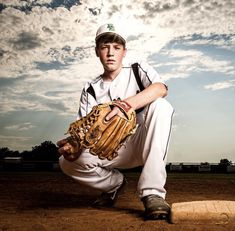 a baseball player kneeling on the ground with his mitt in front of him and clouds behind him