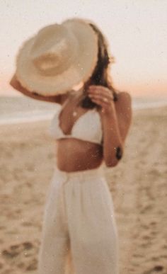 a woman standing on top of a beach next to the ocean holding a white hat