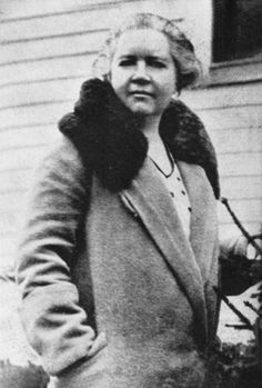 an old black and white photo of a woman standing in front of a house wearing a coat