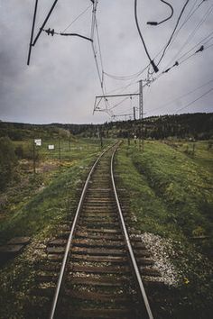 a train track with power lines above it and grass on the other side, under a cloudy sky