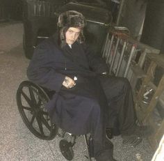 a man sitting in a wheel chair wearing a fur hat and black coat with long sleeves