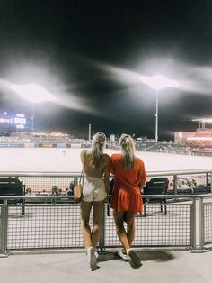 two women are sitting on a fence watching the baseball game at night, with stadium lights in the background