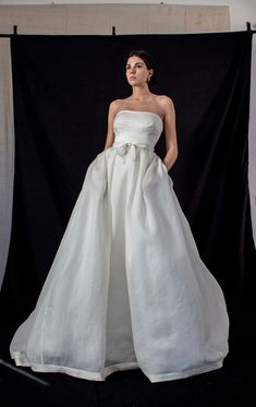 a woman standing in front of a black backdrop wearing a white dress with a bow at the waist