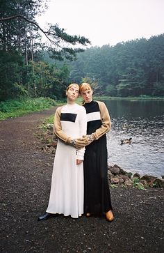 two women standing next to each other on a dirt road near the water and trees