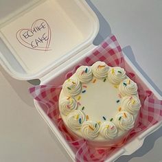 a white cake in a pink and white checkered box with a heart shaped frosting on top