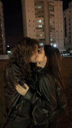 a man and woman are kissing in front of tall buildings at night with their faces close to each other