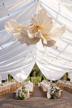 Elevate your outdoor wedding with this stunning setup featuring billowing white drapery, natural wooden seating, and lush floral arrangements. The oversized hanging flower adds a touch of artistic whimsy, creating a breathtaking and romantic atmosphere. Perfect for couples seeking timeless elegance with a modern twist. Explore more wedding decor ideas with The Bridal Journey!