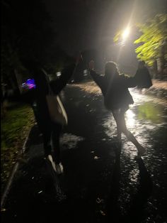 two people walking down a street at night with their arms in the air and one person holding an umbrella