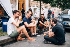 four men are sitting on the curb talking and drinking beer while one man sits in front of them