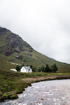 a small white house sitting on the side of a lush green hillside next to a river