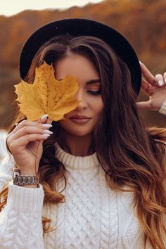 a woman holding a leaf in front of her face while wearing a hat and sweater