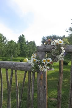 a wreath with daisies and other flowers hangs on a wooden fence in front of a grassy field