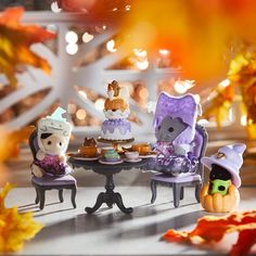 three small figurines sitting at a table with cakes on it and fall leaves in the background