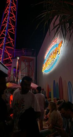 people are sitting outside at night under the neon sign for an amusement park and surf shop